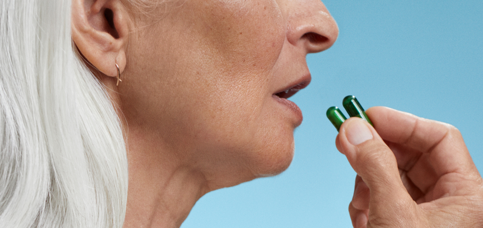 Close-up of a person holding two green capsules near their mouth against a blue background.